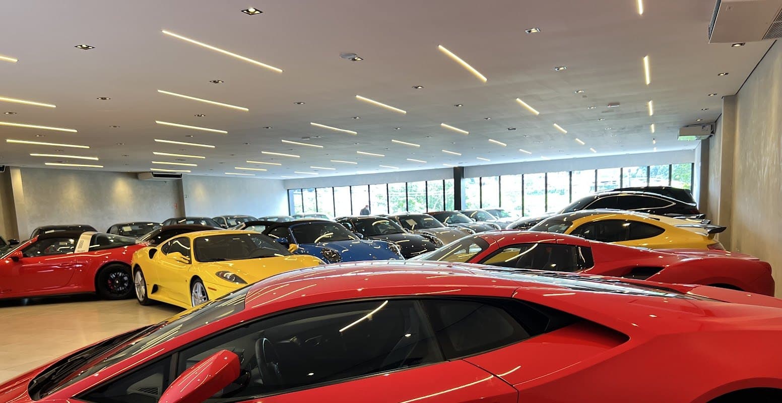 O salão da Castelo Motors por dentro, com uma Lamborghini vermelha em primeiro plano e Ferrari, Porsche e outros esportivos enfileirados sob as fitas de LED do teto